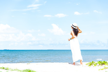 Fototapeta na wymiar Back side of women in white dress and hat walking along the beach with blue sea and sky front view. Travel and summer conceptual.