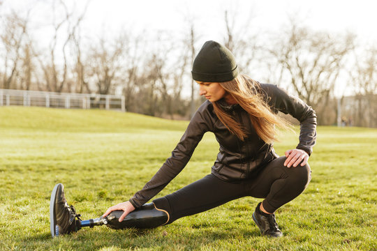 Image of brunette disabled athletic girl in sportswear, doing sports and stretching prosthetic leg on grass