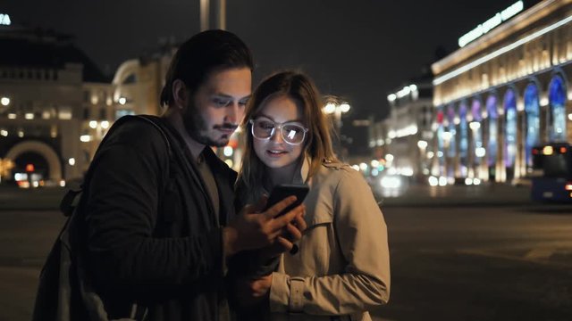 two young female male friends chatting and looking at their smartphone at street night, man and woman happy student tourists