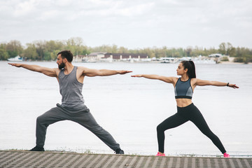 Fototapeta premium Beautiful picture of guy and girl stretching at the river shore. They are stretching and looking to the left. This people are keeping their hands in a horizontal position towards the bridge.