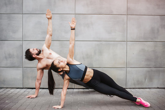 A picture of couple exercising together. They are standing on one hand and reaching up with the other one. They are looking up as well. Couple is concentrated on the exercise.