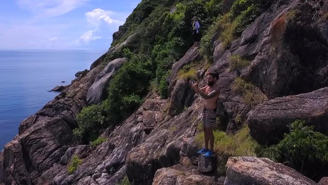 Hikers standing on a rock,admiring beautiful summer sea view, a man makes a photo of the rocky coast along the sea