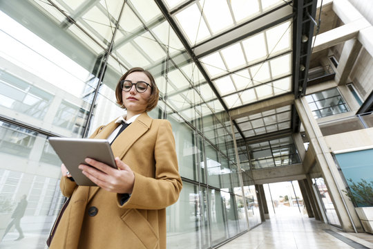 Donna d'affari con occhiali da vista vestita con cappotto beige utilizza il suo tablet in un contesto esterno aziendale 