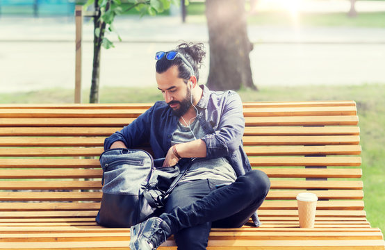 travel, tourism, lifestyle and people concept - man with earphones and coffee sitting on city bench and looking for something in his backpack