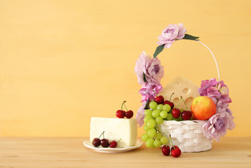 image of fruits and cheese in decorative basket with flowers over wooden table. Symbols of jewish holiday - Shavuot.