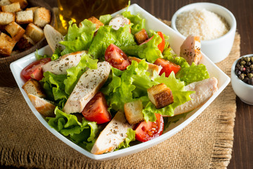 Fresh salad made of tomato, ruccola, chicken breast, eggs, arugula, crackers and spices. Caesar salad in a white, transparent bowl on wooden background