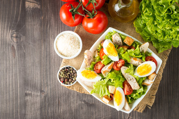 Fresh salad made of tomato, ruccola, chicken breast, eggs, arugula, crackers and spices. Caesar salad in a white, transparent bowl on wooden background