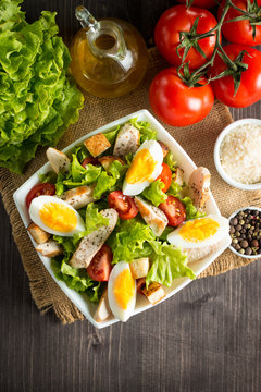 Fresh salad made of tomato, ruccola, chicken breast, eggs, arugula, crackers and spices. Caesar salad in a white, transparent bowl on wooden background