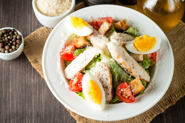 Fresh salad made of tomato, ruccola, chicken breast, eggs, arugula, crackers and spices. Caesar salad in a white, transparent bowl on wooden background