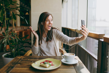 Young woman sitting alone in coffee shop at table with cup of cappuccino, cake, relaxing in restaurant during free time. Young female doing selfie on mobile phone, rest in cafe. Lifestyle concept.