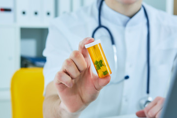 Male medicine doctor hand hold jar of pills and explain how to take pills to patient at worktable. Panacea and life save prescribing treatment legal drug store concept.