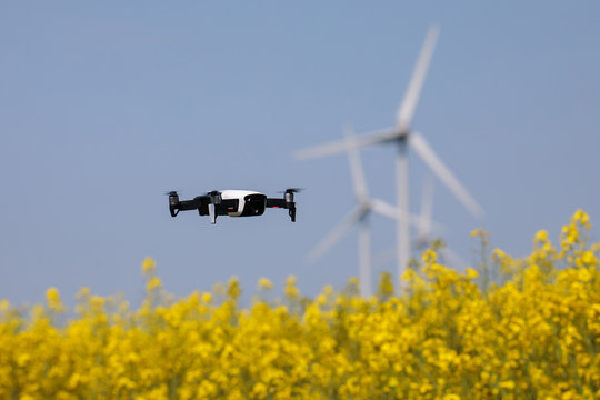 Mini drone floating above rapeseed field surveilling the wind turbines, recording videos and taking pictures. New and modern guardian of big companies. Blurred background