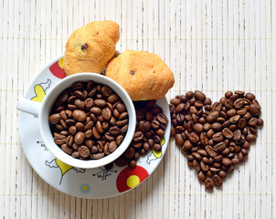 The Cup is filled with coffee beans. On a saucer are croissants. Coffee beans are scattered on the table. Heart of coffee beans.