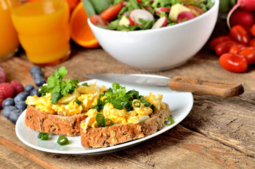 Wholemeal bread with scrambled eggs, fresh herbs, spring onions, orange juice, tomatoes and salad bowl in background