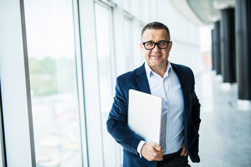 Fototapeta premium Professional at work. Confident senior man in suit and eyeglasses working on laptop while standing in office