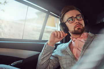 Successful stylish young businessman in a gray suit and pink shirt, riding on a back seat in a luxury car.