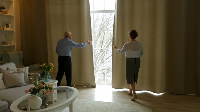 Elderly couple opened the curtains in the living room, embracing and looks out the window