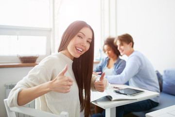 Obraz premium Nice picture of asian girl showing big thumb up. She looks happy. Also there are another people sitting at the table. They are studying.