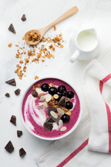 Pink black currant berries, banana smoothie bowl with granola, flakes from almonds and chocolate on light gray concrete background. Selective focus. Top view. Copy space.