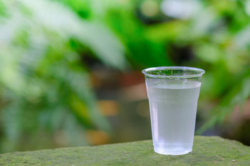 Close up of plastic cup of cold water on stone in the garden with green leaves as background