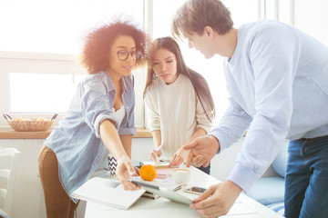 Girl that wears glasses and a guy standing and holding a tablet together while their friend is standing as well and trying to find somthing in her notebook. The asian girl looks serious and