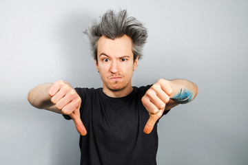 Unshorn and unshaven young guy shows thumbs down, on gray background.