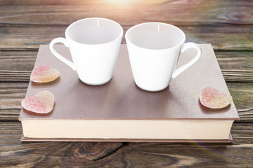 couple mugs, marmalade in the form of hearts on a book on a wooden background. breakfast with two.