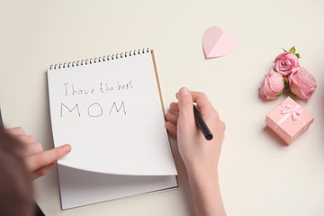Obraz premium Little child writing congratulation for Mother's Day in notebook on light background, top view
