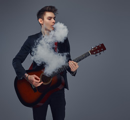 Handsome young musician with stylish hair in elegant clothes exhales smoke while playing acoustic guitar.