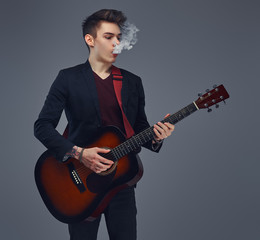 Obraz premium Handsome young musician with stylish hair in elegant clothes exhales smoke while playing acoustic guitar.