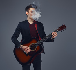 Obraz premium Handsome young musician with stylish hair in elegant clothes exhales smoke while playing acoustic guitar.