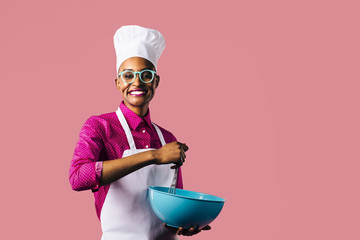 Portrait of a very happy and smiling young woman in cooking hat and apron mixing a bowl with a whisker, isolated on pink studio background  - Powered by Adobe