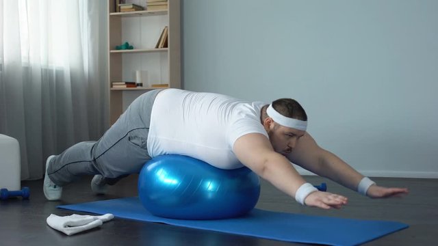 Plump male doing static exercise effort on fitness ball, desire to lose weight
