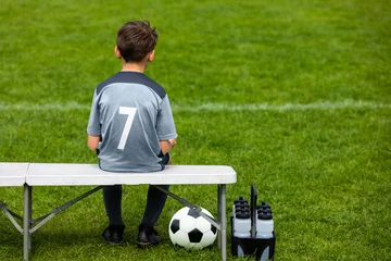 Abwaschbare Fototapete Fußball Little footballer sitting on a wooden bench and watching soccer game. Young substitute player waiting on a soccer bench. Boy on a grass pitch with a soccer ball and water bottles.
