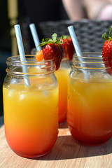 Three summer alcoholic cocktails on a wooden table in the sunlight. Orange and red in colour with fresh strawberry and orange segment on top of glass. Straw in beaker. 