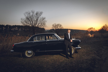 Young handsome groom is next near to black retro car sunset background. Wedding with vintage old...