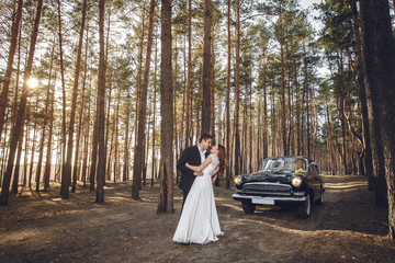 Just married wedding couple is standing near the retro vintage car in the park. Spring or autumn sunny day in forest. bride in elegant white dress with bouquet and elegant groom in love hugging