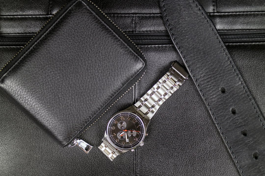 Mens stylish accessories of black leather, wallet, belt and a steel watch on the background of a black briefcase.