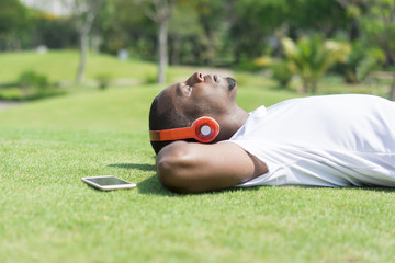 Serene black man resting in park and listening to music. Young Afro American lying on grass with cellphone and colorful wireless headphones. Leisure and connection concept