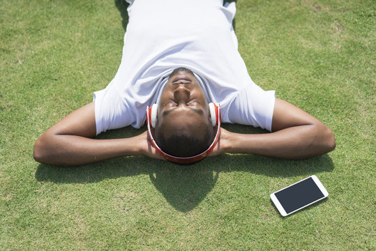 Relaxed black man resting in park and listening to music. Young sporty Afro American lying on grass with wireless headphones and phone. Leisure and technology concept - Powered by Adobe