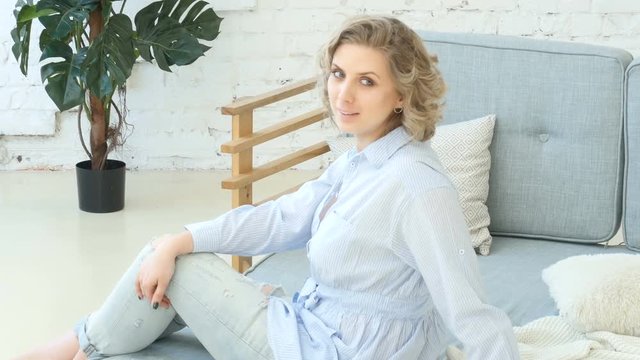Young blonde woman sitting on sofa and posing for photos, girl in casual clothes sits on sofa