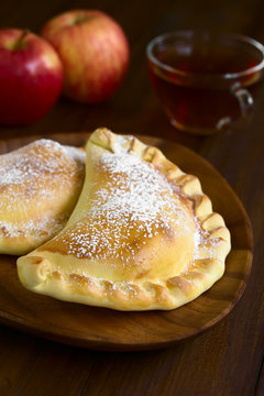 Chilean Apple Empanada With Icing Sugar, Cup Of Tea In The Back, Photographed On Dark Wood With Natural Light (Selective Focus, Focus One Third Into The Image)