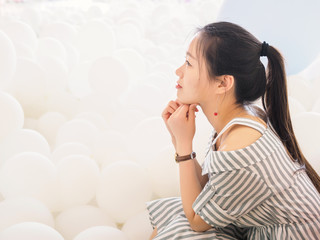 Portrait of beautiful Chinese girl in dress sit among white balloons with hands on chin, Beautiful Happy Young woman on birthday holiday party to make a wish.