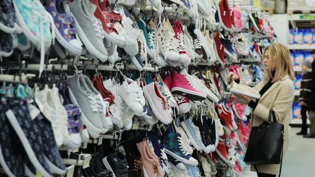 calm beautiful girl holding a pair of stylish sneakers while standing near a shelf of shoes in supermarket shopping mall warehouse blonde woman choosing new summer shoes in shoe store clothes business