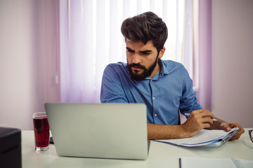 Young student with a beard is making notes and working with laptop