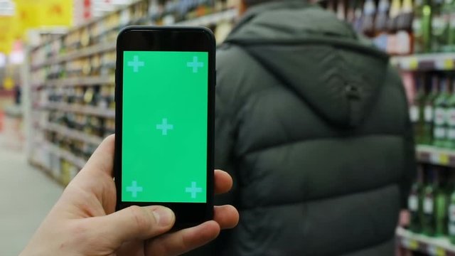 closeup view of man hand holding and using modern smartphone mobile with empty green screen in supermarket and shelves with alcohol drinks on blurred background tapping chromakey mock-up free content
