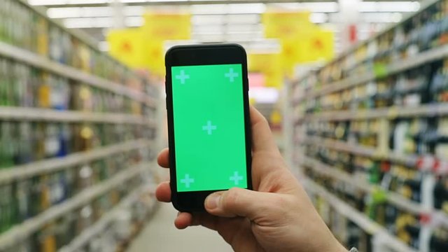 close-up view of man hand holding modern smartphone mobile with empty green screen in supermarket and shelves with bottles of alcohol beverages on blurred background chromakey mockup free content