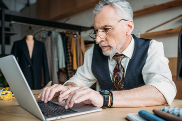 close-up portrait of handsome mature tailor working with laptop at sewing workshop