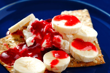 Close up dessert with dietary cookies, banana, raspberry topping and cream cheese mascarpone in blue plate on white wooden table. Healthy food. Soft focus