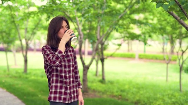 Young pretty girl standing on path in the park and drinking a cup of coffee